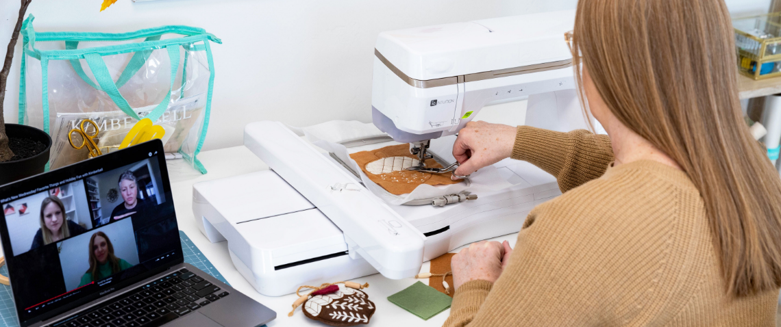 Woman stitching acorn stuffie on embroidery machine while on a video call with other event attendees. 